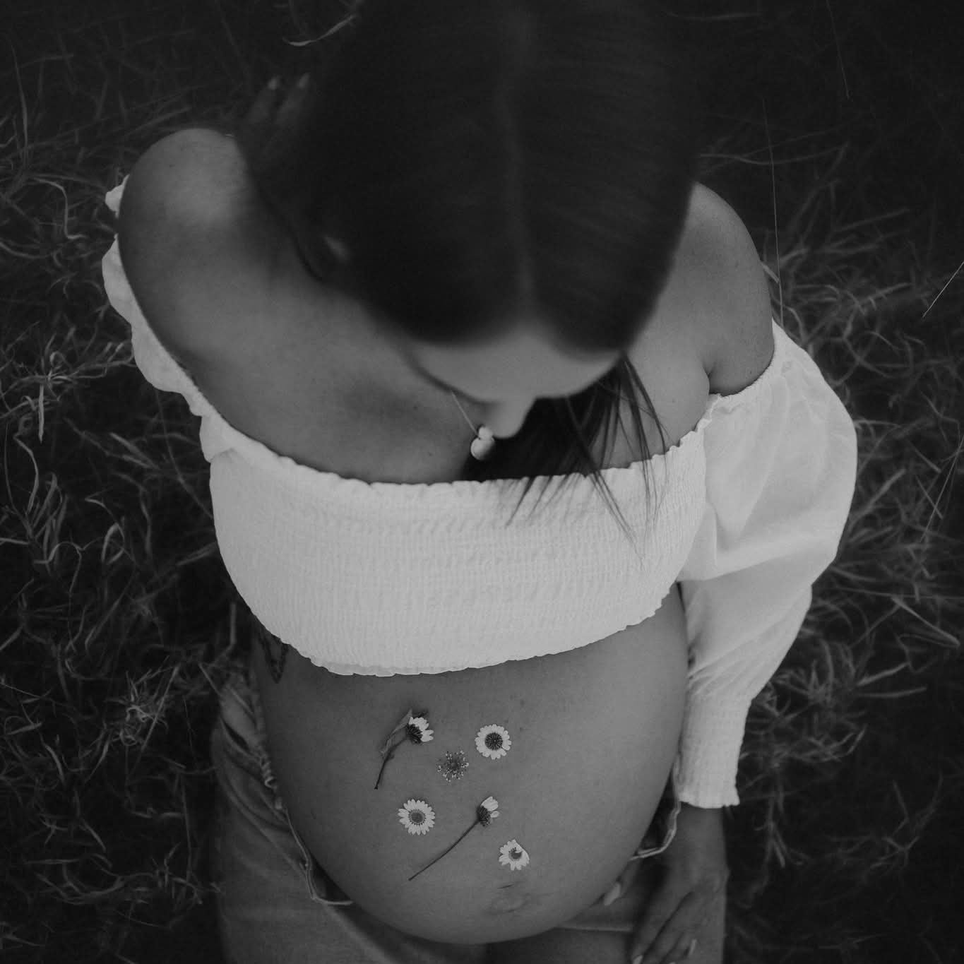 Black and white photo of a pregnant person with daisy flowers on their belly in a field.